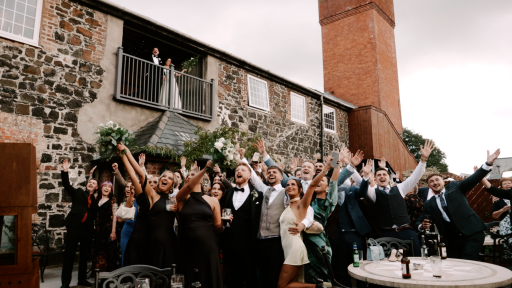 All the guests cheering for a group photo outside The Wool Tower Ballymena while the bride and groom cheer from a balcony above.